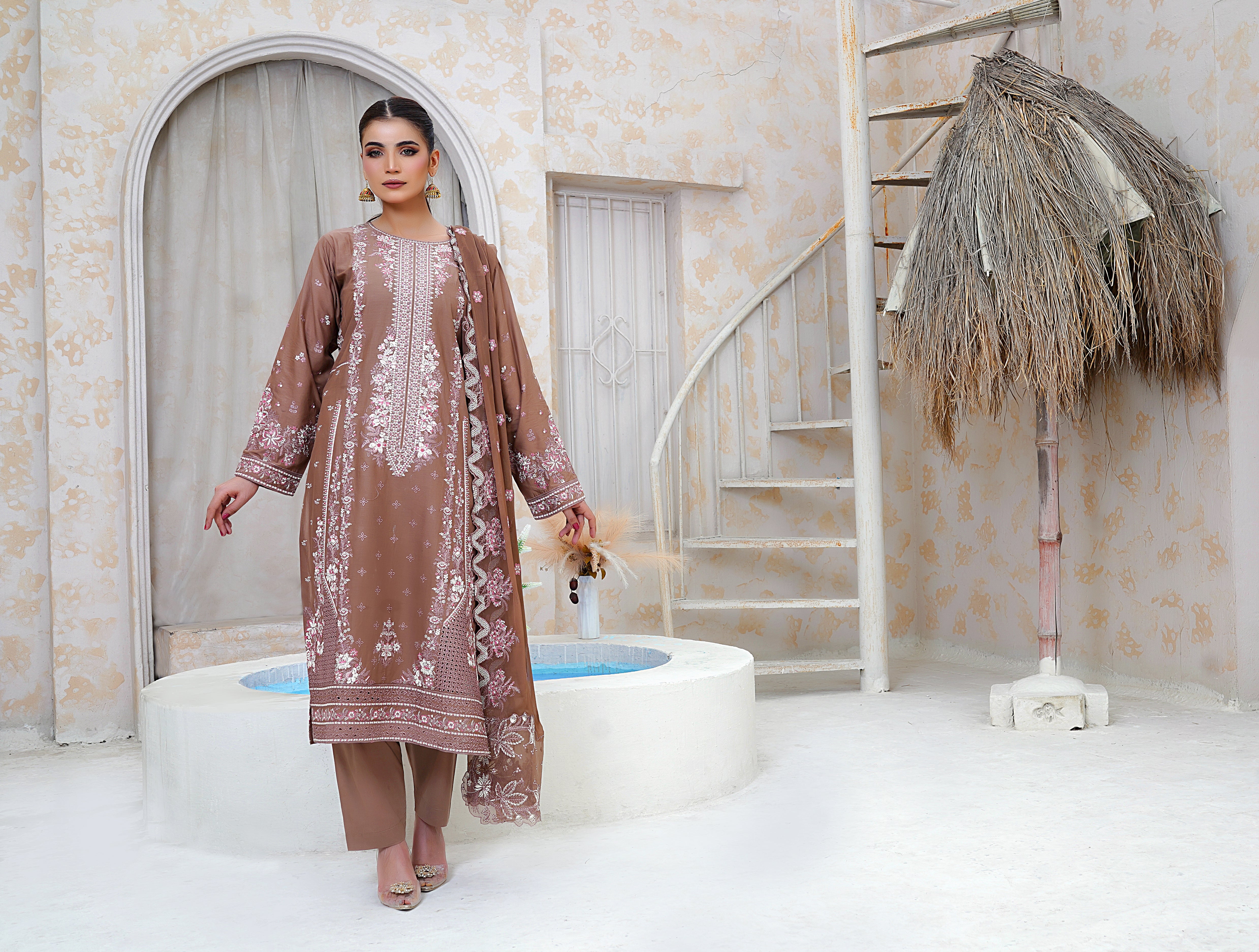 Woman in a brown embroidered outfit standing in a decorative room with a staircase and decorative elements.